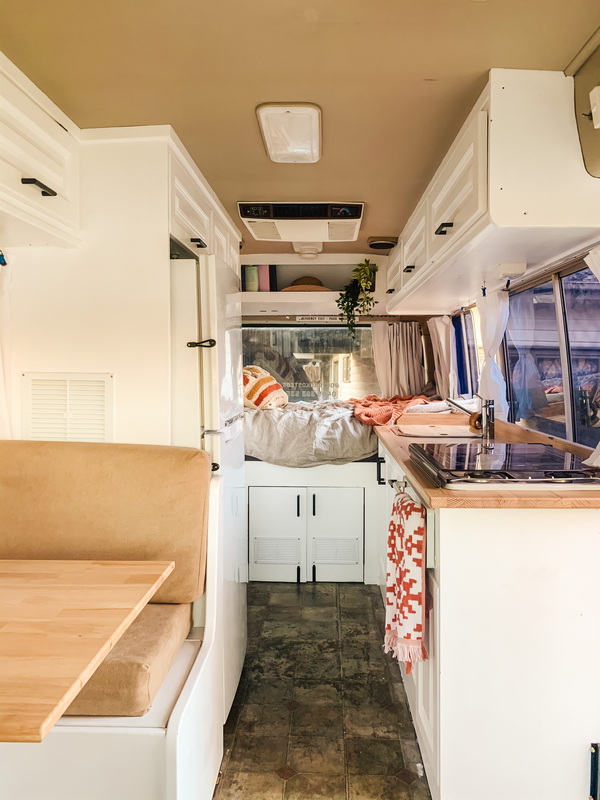 Interior of modernised campervan with white cupboard, looking from front past table, kitchen and to an unmade bed.