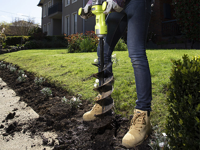 A person using a RYOBI planting and digging tool to dig holes into a garden bed in a front yard, ready for planting. 
