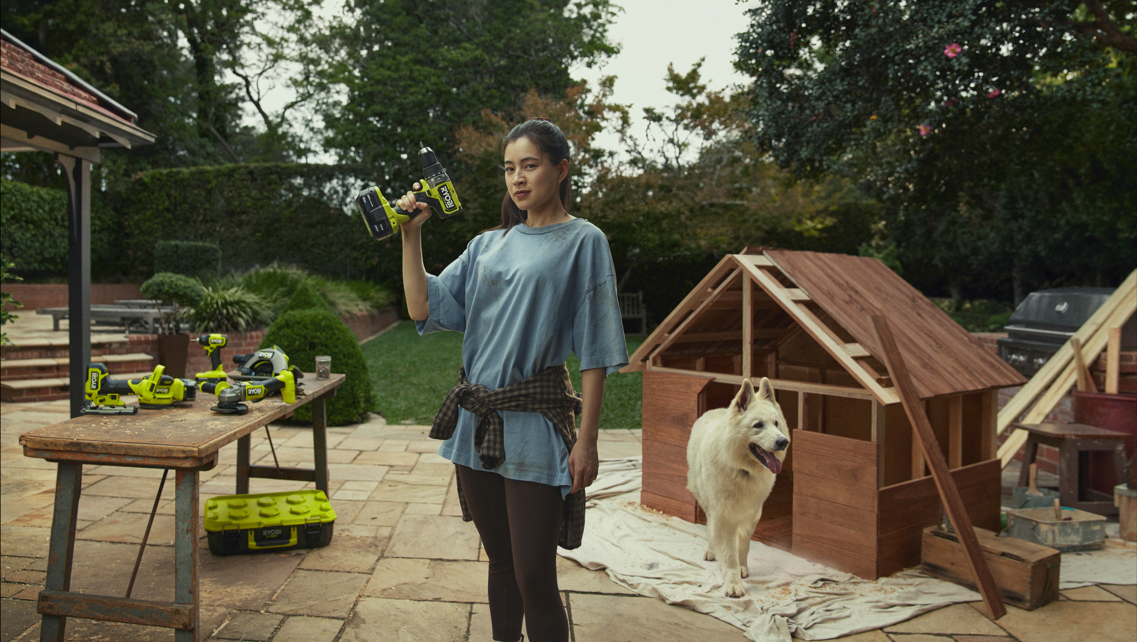 A woman stands holding a RYOBI Drill. Behind her is a DIY kennel, a fluffy white dog and a table full of RYOBI power tools.