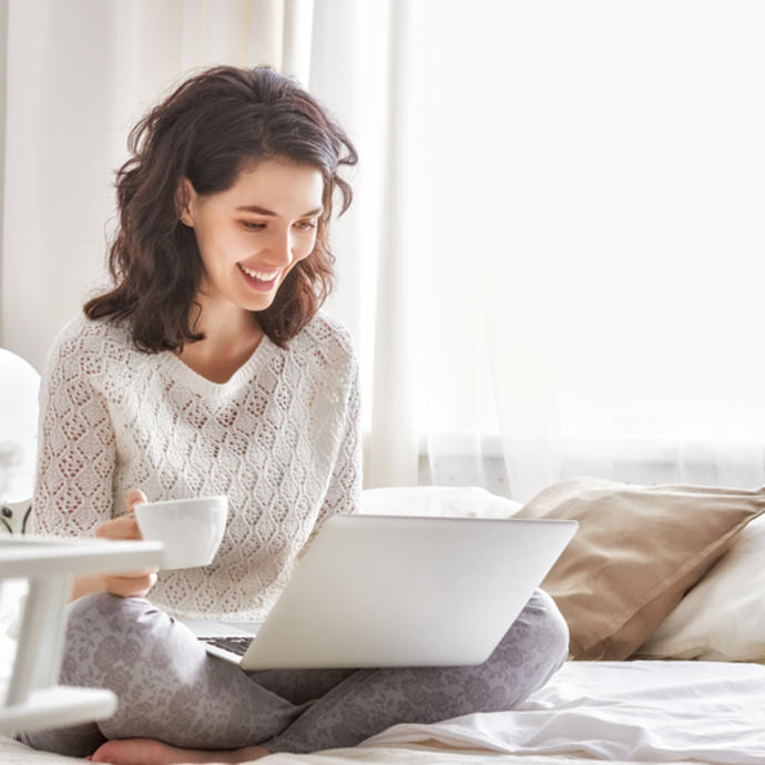 A woman sits, working on her laptop holding a cup of tea with a large smile. The room around her is clean and tidy.